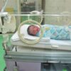 A newborn baby lies in an incubator at a hospital, wrapped in a colorful blanket.