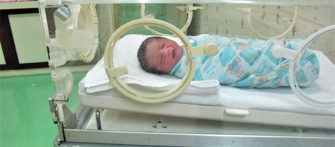 A newborn baby lies in an incubator at a hospital, wrapped in a colorful blanket.