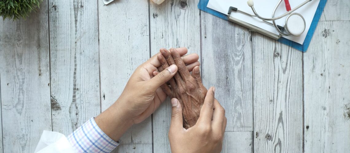 A doctor holding an elderly patient's hands on a desk with medication and stethoscope.