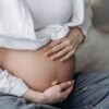 Close-up of a pregnant woman sitting, cradling her bare belly with both hands.