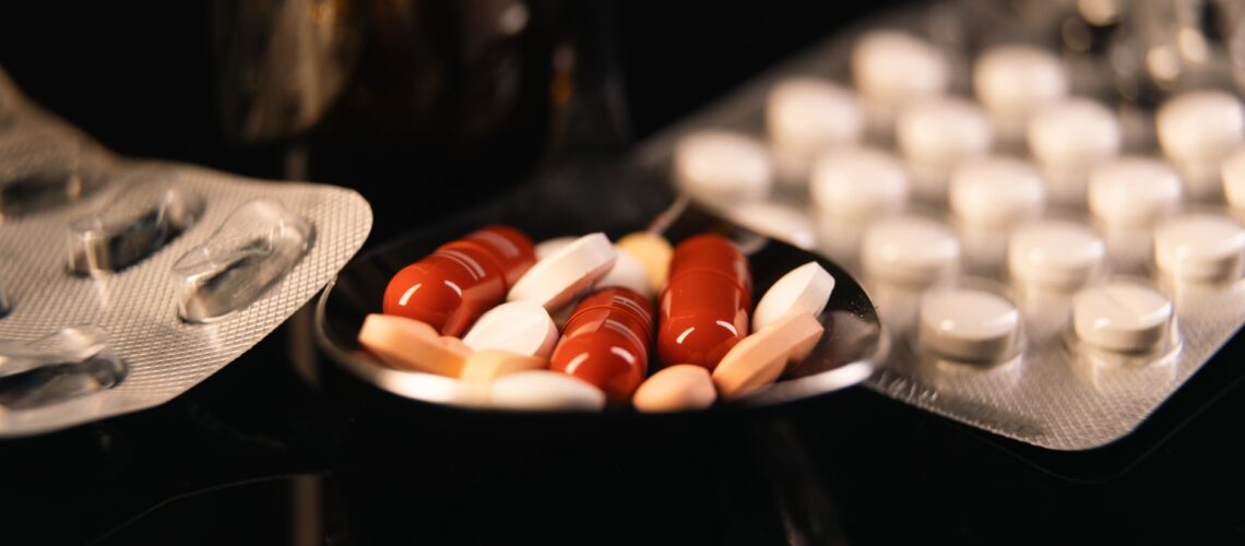 Close-up of assorted pharmaceutical pills in blister packs and a spoon on a dark background.