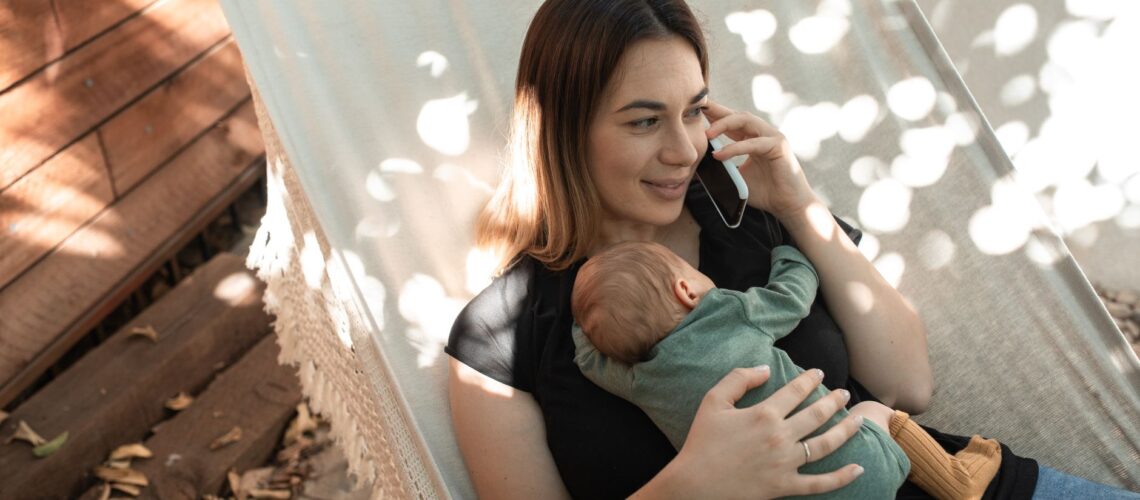 Mother talking on phone while holding baby in a hammock outdoors.