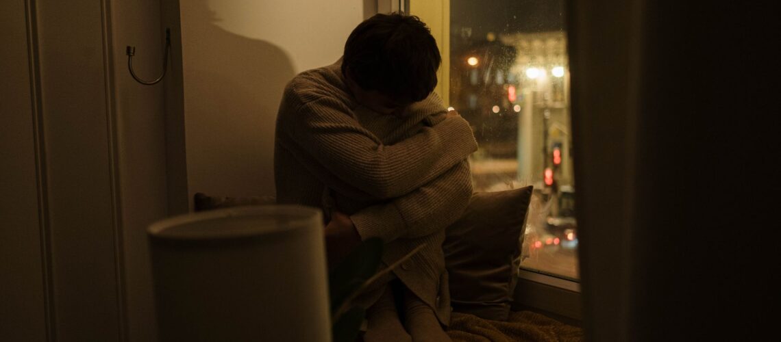 A woman sits alone by a window at night, depicting solitude and reflection.
