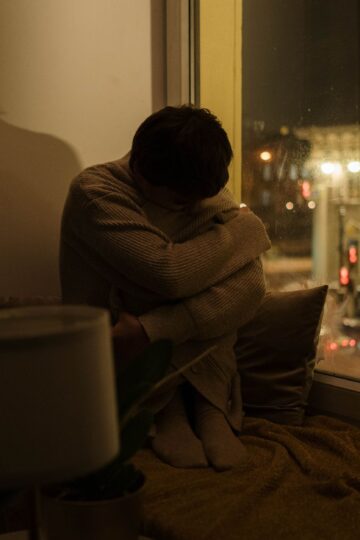 A woman sits alone by a window at night, depicting solitude and reflection.