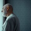 A senior man with a beard looks thoughtfully out of a sunny window, reflecting on life indoors.