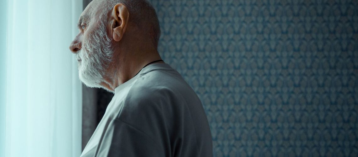 A senior man with a beard looks thoughtfully out of a sunny window, reflecting on life indoors.