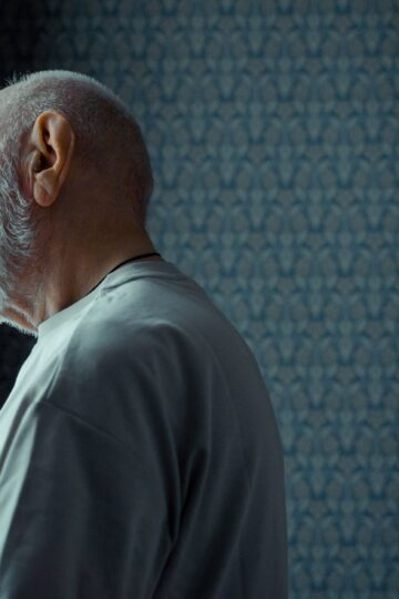 A senior man with a beard looks thoughtfully out of a sunny window, reflecting on life indoors.