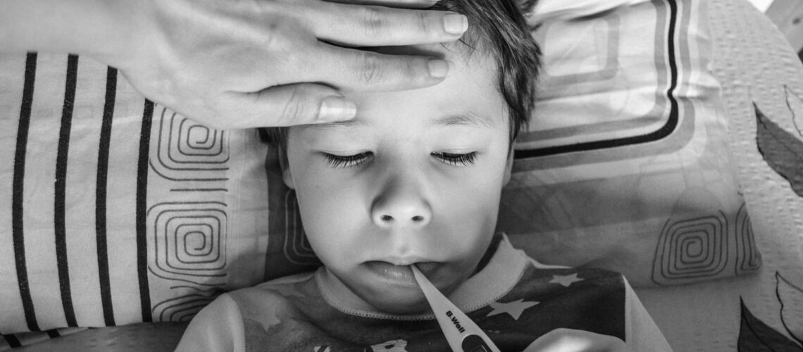 Black and white from above of unrecognizable person touching forehead of unemotional child in casual clothing holding electronic thermometer in mouth