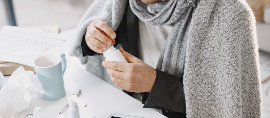 Adult male in scarf taking medicine at a desk with tissues and tablet, feeling unwell.