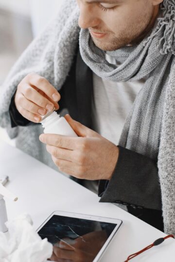Adult male in scarf taking medicine at a desk with tissues and tablet, feeling unwell.