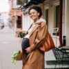 A smiling pregnant woman in a brown coat holding a bouquet on a vibrant city street.