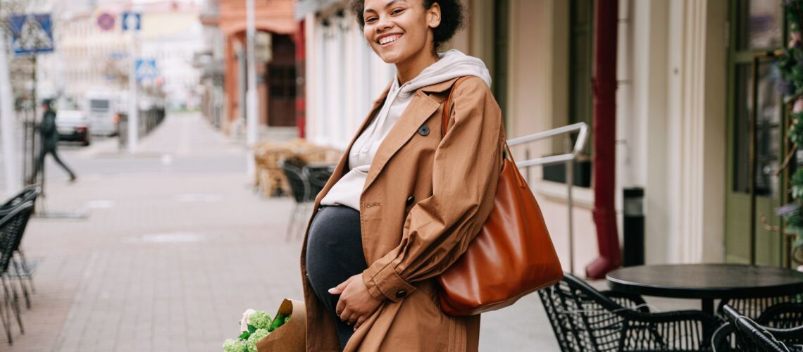 A smiling pregnant woman in a brown coat holding a bouquet on a vibrant city street.