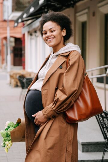 A smiling pregnant woman in a brown coat holding a bouquet on a vibrant city street.
