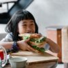 Asian child sitting at wooden table during breakfast and eating tasty sandwich with lettuce leaves