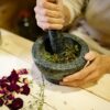A person grinding herbal ingredients in a stone mortar and pestle with dried petals nearby.