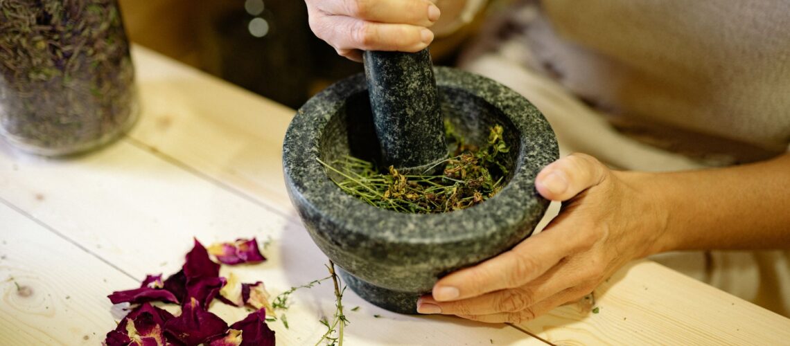 A person grinding herbal ingredients in a stone mortar and pestle with dried petals nearby.