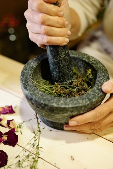 A person grinding herbal ingredients in a stone mortar and pestle with dried petals nearby.