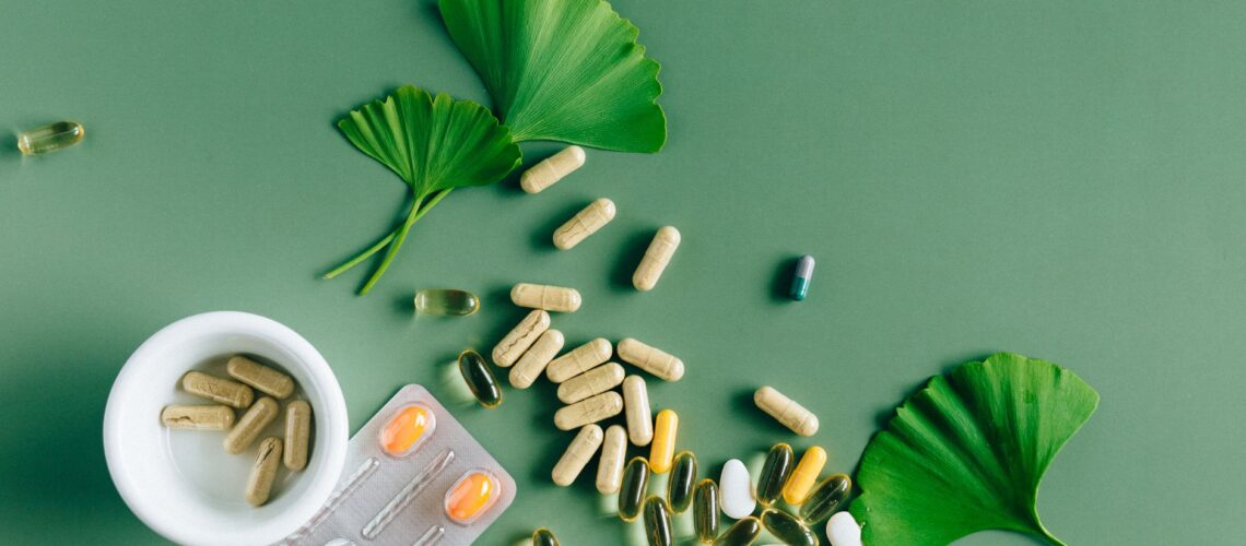 Flat lay of varied supplements and medicines with ginkgo leaves on green surface, showcasing a natural remedy concept.