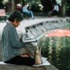 Elderly woman enjoying a peaceful reading moment by a pond in a serene park setting.
