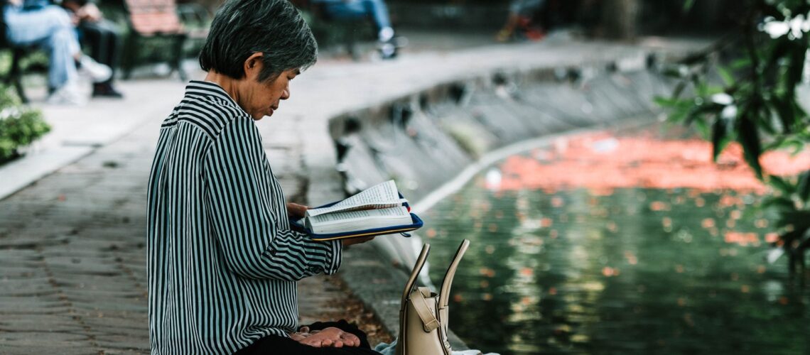 Elderly woman enjoying a peaceful reading moment by a pond in a serene park setting.