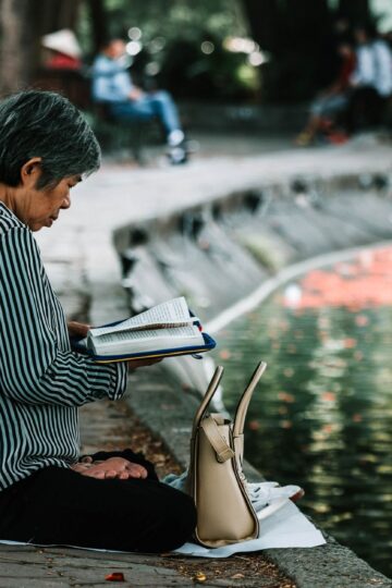 Elderly woman enjoying a peaceful reading moment by a pond in a serene park setting.