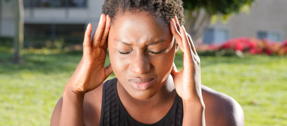 A woman holding her head in pain, indicating a severe headache outdoors in a sunny setting.