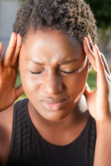 A woman holding her head in pain, indicating a severe headache outdoors in a sunny setting.