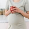 Unrecognizable pregnant female with ripe red apple for healthy diet in hands standing near counter in light kitchen at home