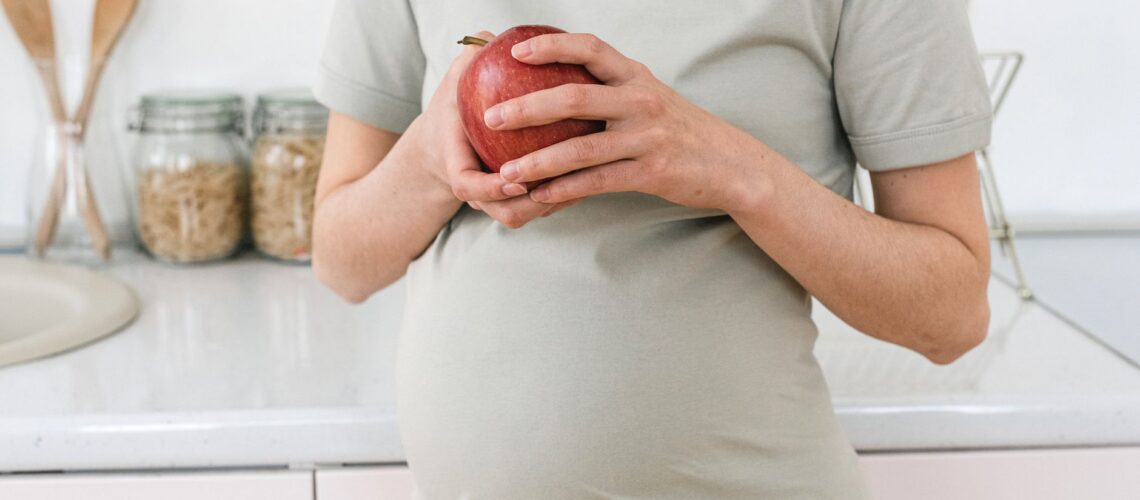 Unrecognizable pregnant female with ripe red apple for healthy diet in hands standing near counter in light kitchen at home