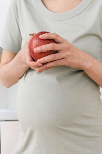 Unrecognizable pregnant female with ripe red apple for healthy diet in hands standing near counter in light kitchen at home
