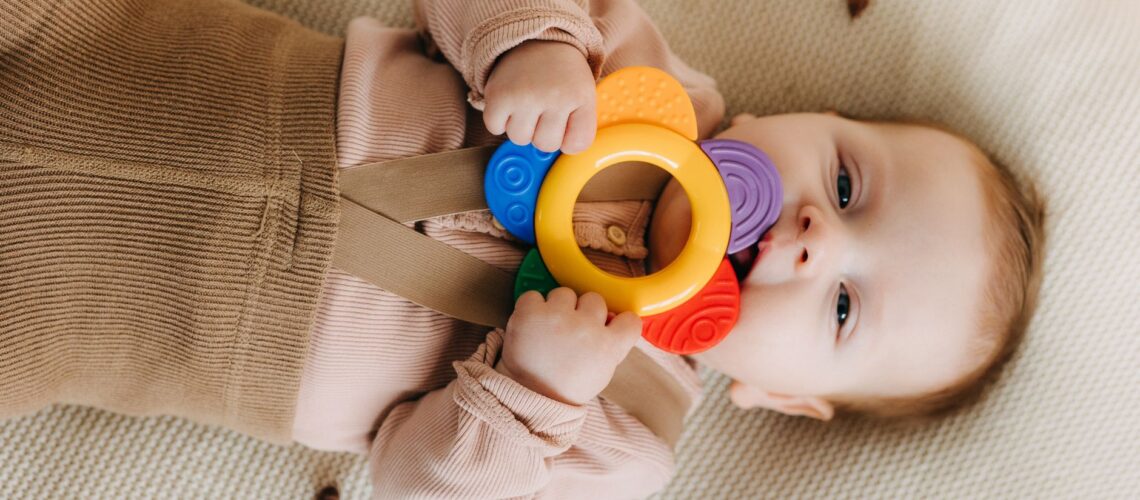 Cute baby chewing on a colorful teething toy while lying on a soft surface.