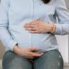 Pregnant woman sitting indoors, hands on belly, wearing casual clothes. Healthcare consultation setting.