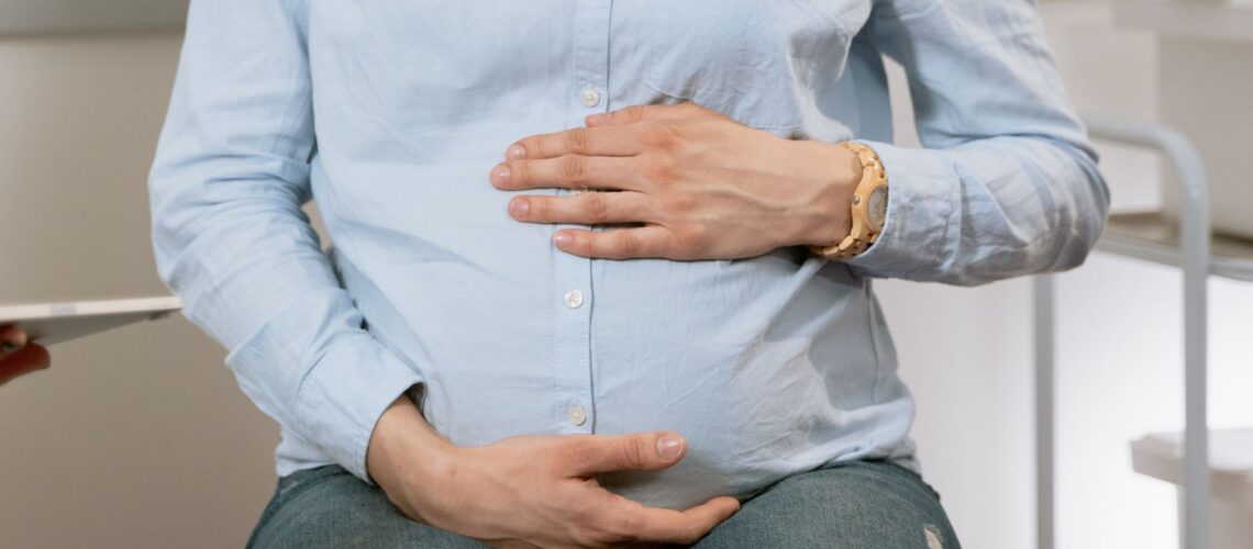 Pregnant woman sitting indoors, hands on belly, wearing casual clothes. Healthcare consultation setting.