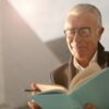 Elderly man wearing glasses, reading a book outdoors with a smile on a sunny day.