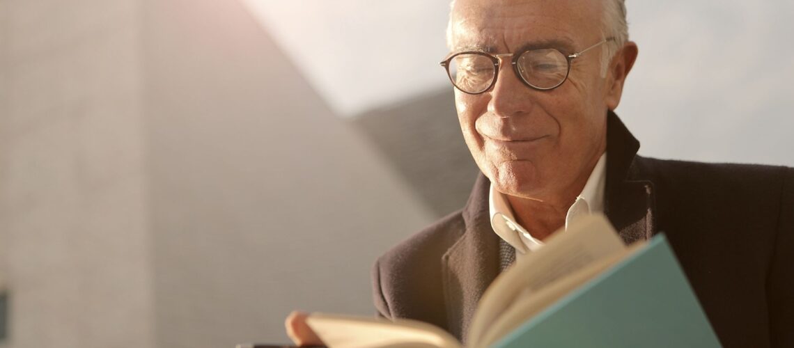 Elderly man wearing glasses, reading a book outdoors with a smile on a sunny day.