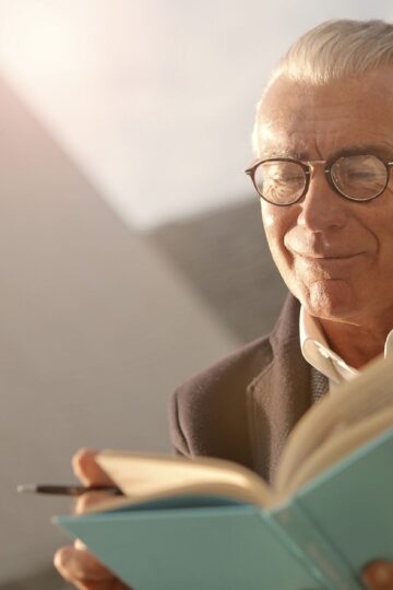 Elderly man wearing glasses, reading a book outdoors with a smile on a sunny day.