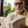 Elderly man with eyeglasses focused on reading a book outside.