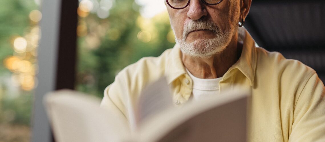 Elderly man with eyeglasses focused on reading a book outside.