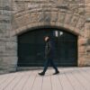 Elderly man walking past a rustic stone building exterior, hands in pockets. Urban scene.
