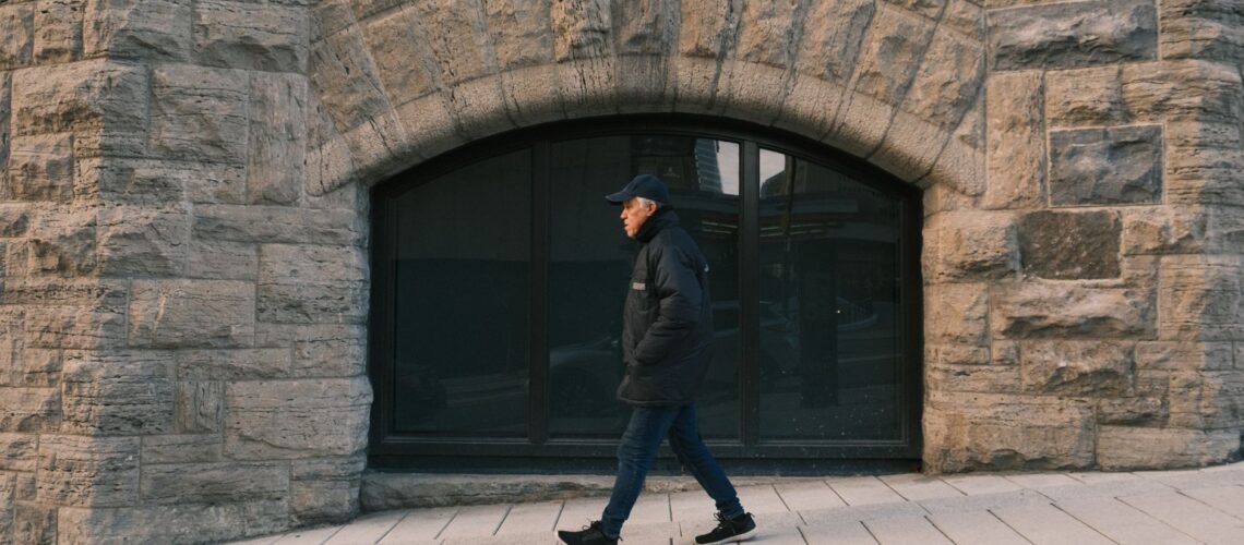 Elderly man walking past a rustic stone building exterior, hands in pockets. Urban scene.