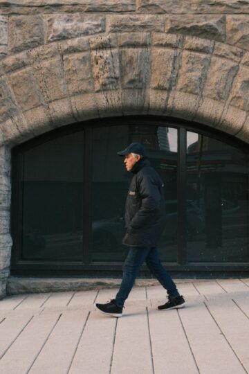 Elderly man walking past a rustic stone building exterior, hands in pockets. Urban scene.