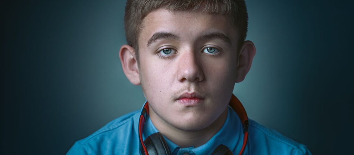 Portrait of a serious teenage boy wearing a blue blouse and headphones against a gray background.