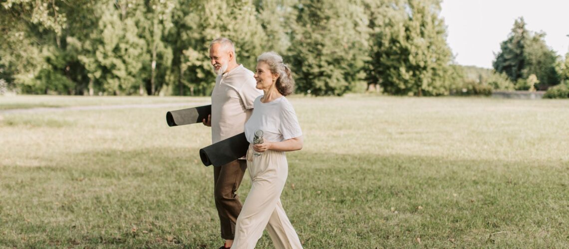 Elderly couple walking on grass carrying yoga mats, enjoying a sunny day outdoors.