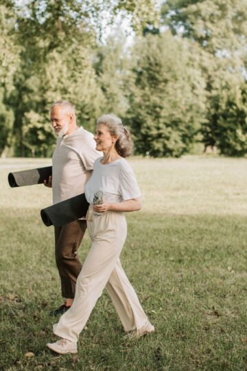 Elderly couple walking on grass carrying yoga mats, enjoying a sunny day outdoors.
