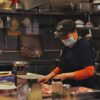 A woman in a kitchen wears a face mask while cutting meat, showcasing food preparation safety.