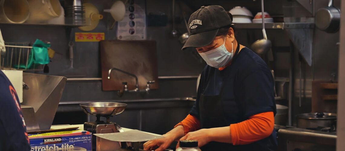 A woman in a kitchen wears a face mask while cutting meat, showcasing food preparation safety.