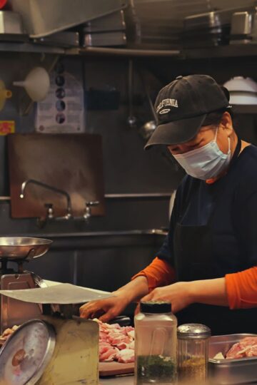 A woman in a kitchen wears a face mask while cutting meat, showcasing food preparation safety.
