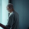 A senior man stands alone, looking out a window with an expression of deep contemplation.