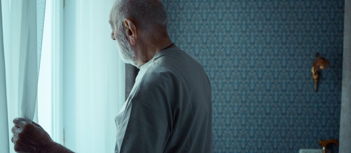 A senior man stands alone, looking out a window with an expression of deep contemplation.