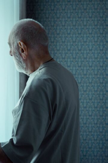 A senior man stands alone, looking out a window with an expression of deep contemplation.